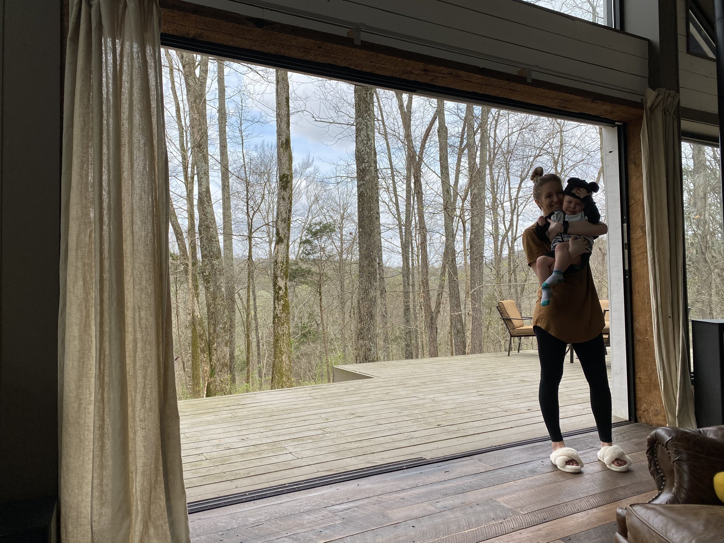 Liz holding her toddler in a doorway opening onto a wooden deck and bare winter woods