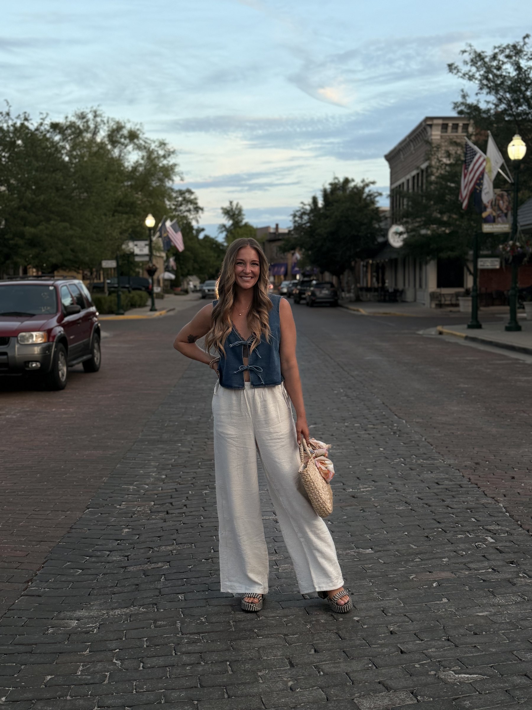 Liz standing on a brick main street at dusk in a denim vest and linen trousers